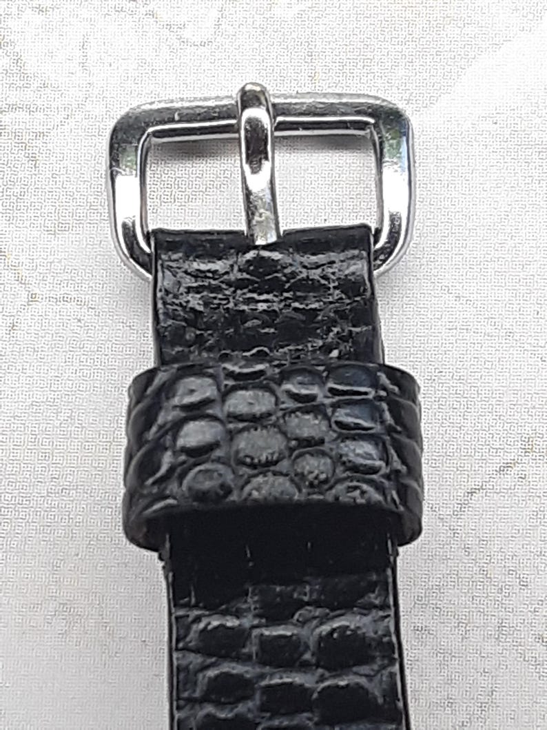 May include: Close-up of a black watch strap with a silver-colored buckle. The strap has a textured, embossed pattern. The buckle is rectangular with a single bar for securing the strap. The watch strap is likely made of leather or a leather-like material.