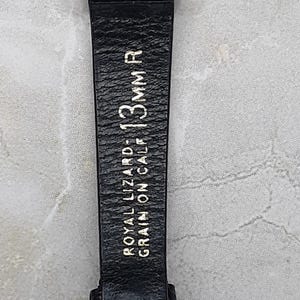 May include: Close-up of a black watch strap with the text "ROYAL LIZARD - GRAIN ON CALF - 13mm R" in gold lettering. The strap is made of textured leather and is attached to a silver watch face.