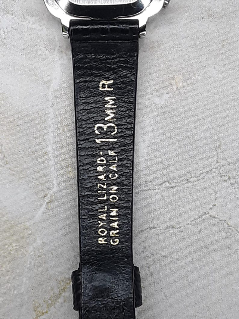 May include: Close-up of a black watch strap with the text "ROYAL LIZARD - GRAIN ON CALF - 13mm R" in gold lettering. The strap is made of textured leather and is attached to a silver watch face.