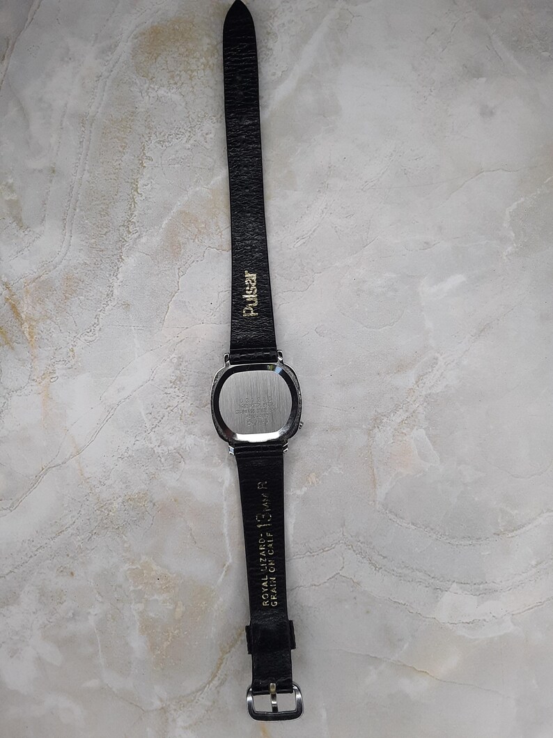 May include: A vintage Pulsar watch with a rectangular silver-tone case and a black leather band. The band is marked with "Pulsar" and "Royal Lizard - 13, AM R Grain on Calf". The watch has a silver buckle.