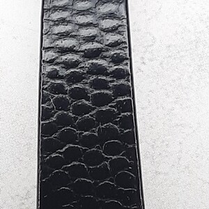 May include: Close-up of a black leather strap with a textured, embossed pattern. The strap appears to be made of genuine leather, with a repeating scale-like design. The strap is rectangular and appears to be a belt or watch band.