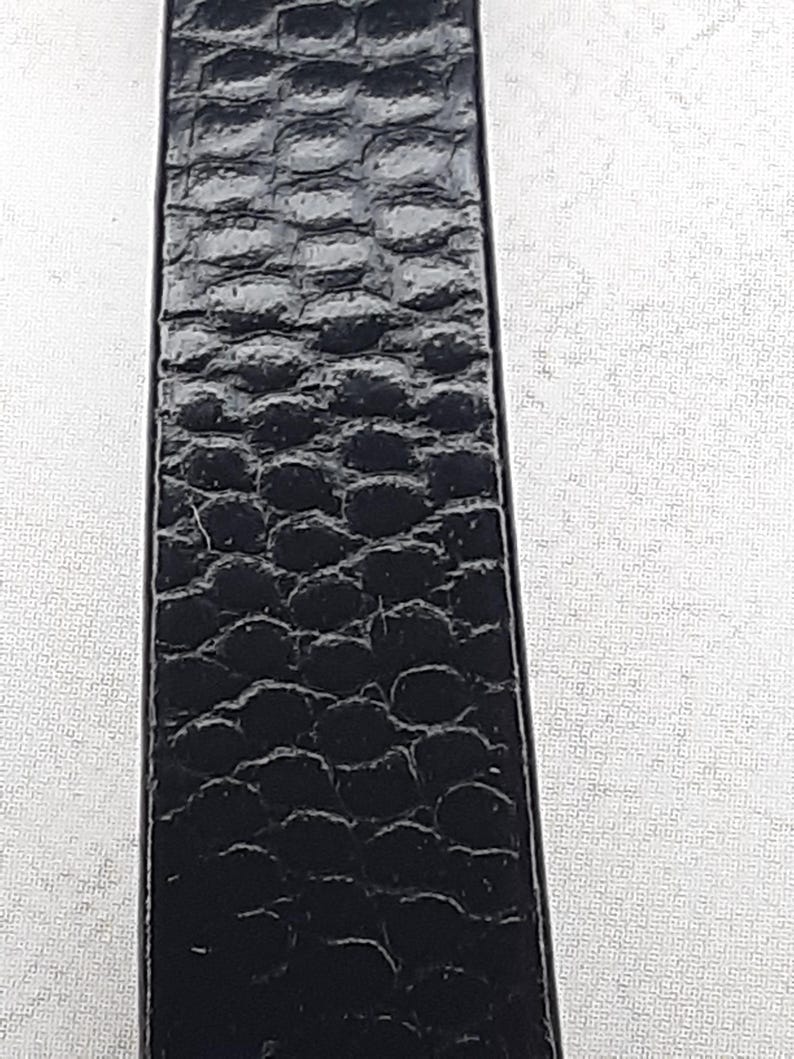 May include: Close-up of a black leather strap with a textured, embossed pattern. The strap appears to be made of genuine leather, with a repeating scale-like design. The strap is rectangular and appears to be a belt or watch band.