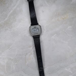 May include: A digital wristwatch with a black textured leather band and a silver-toned square face. The watch displays the time "3:27" and has a silver buckle. The watch is a classic accessory.