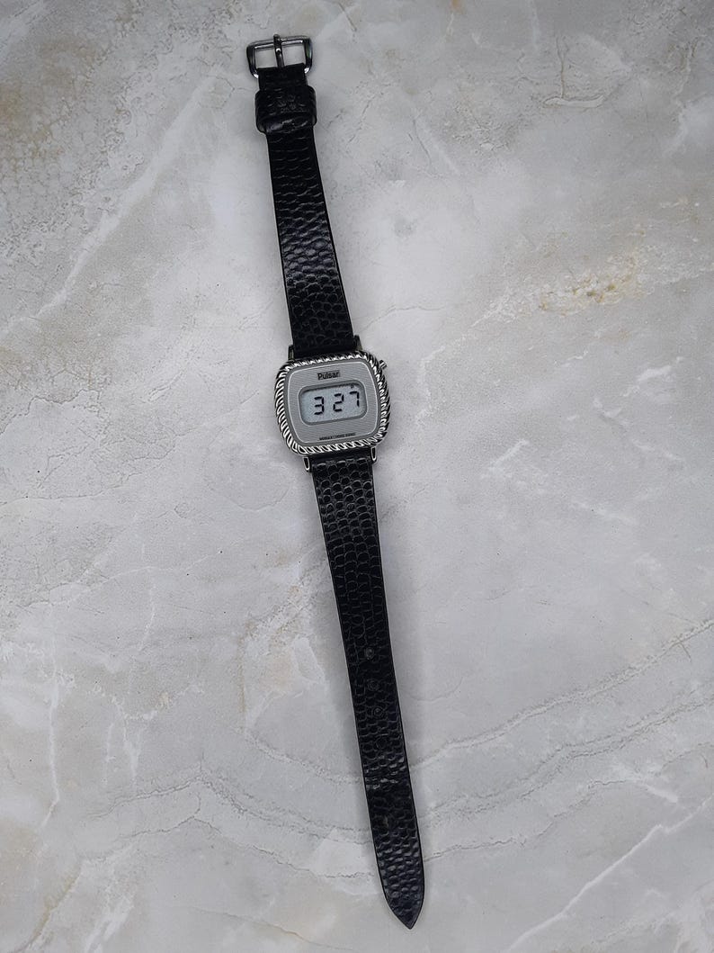 May include: A digital wristwatch with a black textured leather band and a silver-toned square face. The watch displays the time "3:27" and has a silver buckle. The watch is a classic accessory.