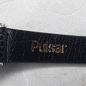 May include: Close-up of a black watch strap with a textured, crocodile-like pattern. The gold-colored word "Pulsar" is embossed on the strap. The watch strap is likely made of leather or a similar material.