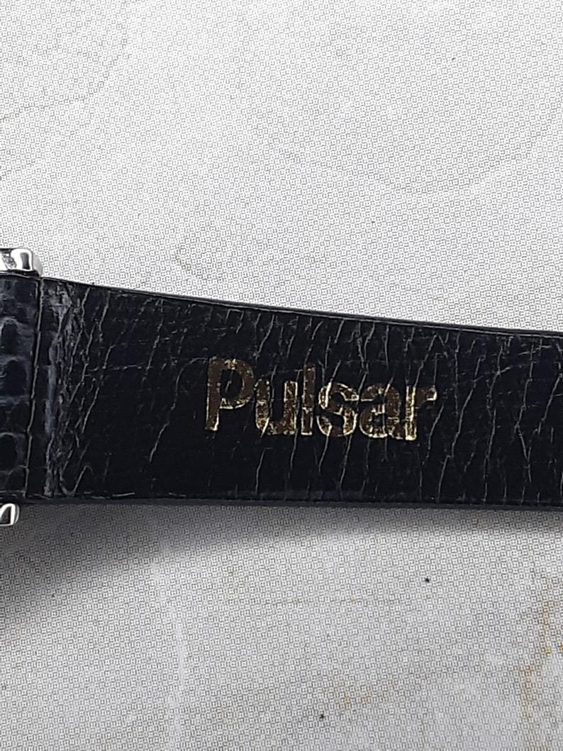 May include: Close-up of a black watch strap with a textured, crocodile-like pattern. The gold-colored word "Pulsar" is embossed on the strap. The watch strap is likely made of leather or a similar material.