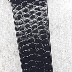May include: Close-up of a black watch strap with a textured, reptile-like pattern. The strap is made of a shiny, dark material and is attached to a silver-colored watch case.