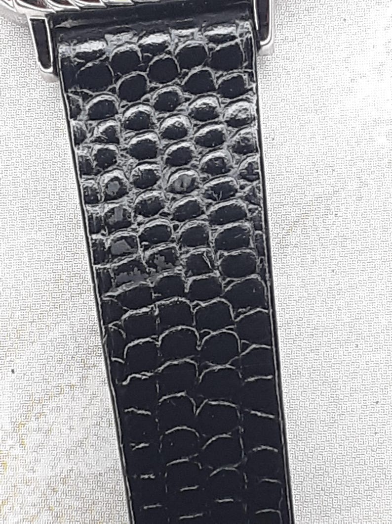 May include: Close-up of a black watch strap with a textured, reptile-like pattern. The strap is made of a shiny, dark material and is attached to a silver-colored watch case.