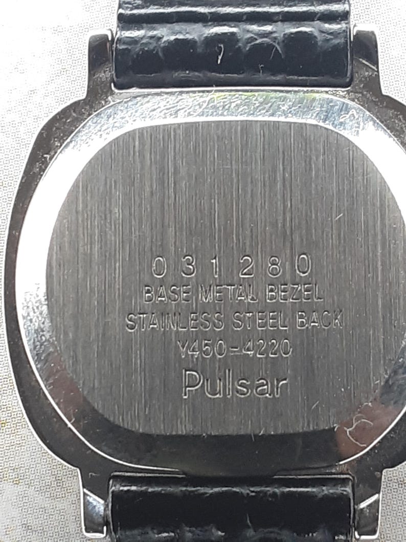 May include: Close-up of a watch back with a silver-colored, brushed metal finish. The watch has a black leather band. The text on the watch reads "031280 BASE METAL BEZEL STAINLESS STEEL BACK Y450-4220 Pulsar."