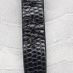 May include: Close-up of a black watch strap with a textured, reptile-skin pattern. The strap has a rectangular shape and is secured with a metal rivet. The background is a light, patterned fabric.