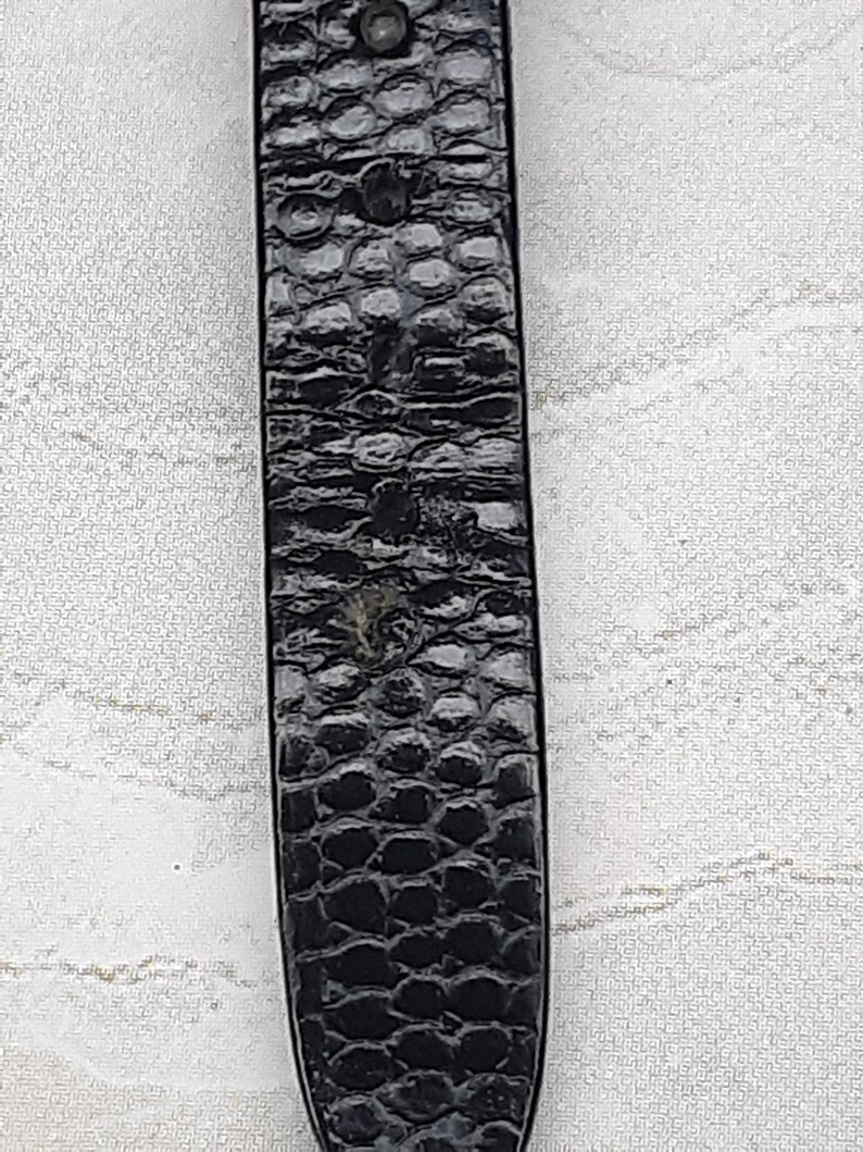 May include: Close-up of a black watch strap with a textured, reptile-skin pattern. The strap has a rectangular shape and is secured with a metal rivet. The background is a light, patterned fabric.