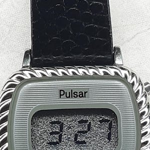 May include: Silver-toned Pulsar digital watch with a black leather band. The watch face displays the time as 3:27. The watch has a rectangular face with a textured silver background and a decorative rope-like border.