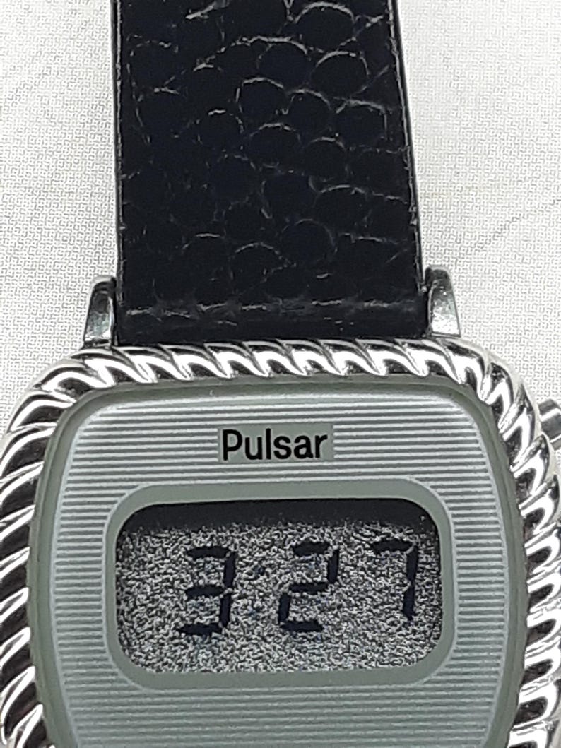 May include: Silver-toned Pulsar digital watch with a black leather band. The watch face displays the time as 3:27. The watch has a rectangular face with a textured silver background and a decorative rope-like border.