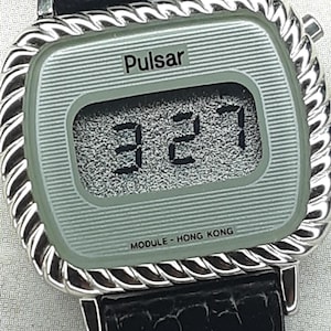 May include: A vintage Pulsar digital watch with a silver-tone case and a black leather band. The watch face displays the time 3:27 in black digits. The watch has a textured, light green face and the text "MODULE - HONG KONG".