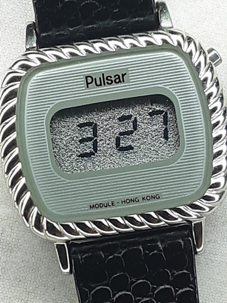 May include: A vintage Pulsar digital watch with a silver-tone case and a black leather band. The watch face displays the time 3:27 in black digits. The watch has a textured, light green face and the text "MODULE - HONG KONG".
