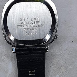 May include: Close-up of a silver-toned watch with a black leather band. The watch face is not visible. The back of the watch has the text "031280 BASE METAL BEZEL STAINLESS STEEL BACK Y450-4220 Pulsar".