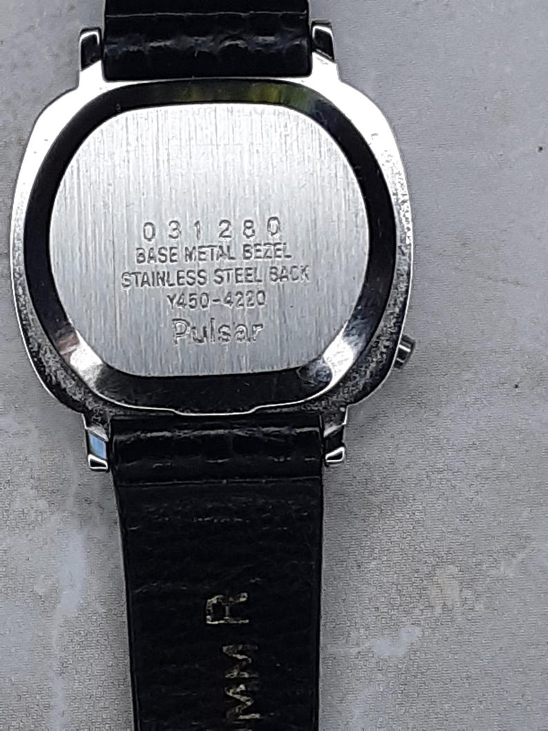 May include: Close-up of a silver-toned watch with a black leather band. The watch face is not visible. The back of the watch has the text "031280 BASE METAL BEZEL STAINLESS STEEL BACK Y450-4220 Pulsar".