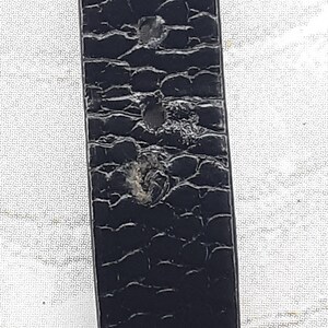 May include: Close-up of a black leather belt with a textured, reptile-like pattern. The belt shows signs of wear, with several small holes and abrasions visible on the surface. The belt is a classic accessory.