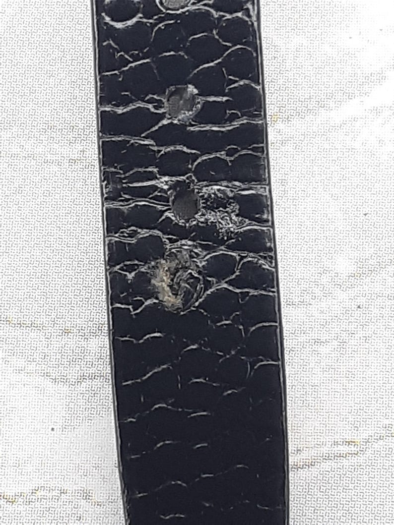 May include: Close-up of a black leather belt with a textured, reptile-like pattern. The belt shows signs of wear, with several small holes and abrasions visible on the surface. The belt is a classic accessory.
