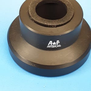 May include: A black metal component with a circular design. The component has a wide base, a raised central section, and a dark, porous ring around the inner opening. The text "A+F SWITZERLAND" is printed on the side.