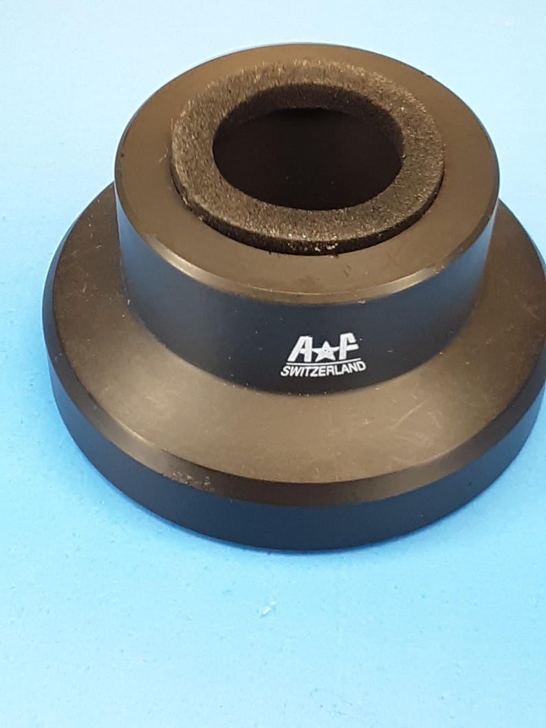 May include: A black metal component with a circular design. The component has a wide base, a raised central section, and a dark, porous ring around the inner opening. The text "A+F SWITZERLAND" is printed on the side.