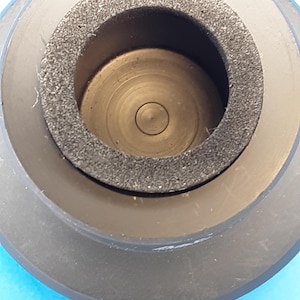 May include: Close-up of a black, cylindrical object with a central opening. The object has a dark gray, textured ring around the opening and a series of concentric circles inside. The object appears to be made of plastic or rubber.
