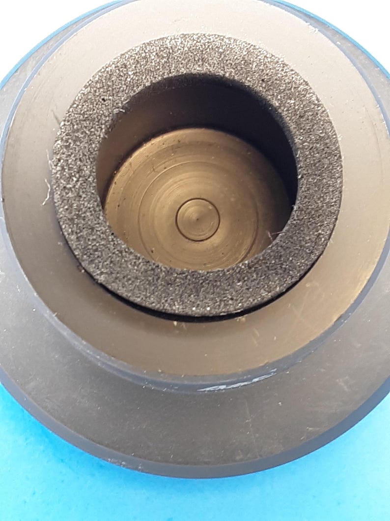 May include: Close-up of a black, cylindrical object with a central opening. The object has a dark gray, textured ring around the opening and a series of concentric circles inside. The object appears to be made of plastic or rubber.