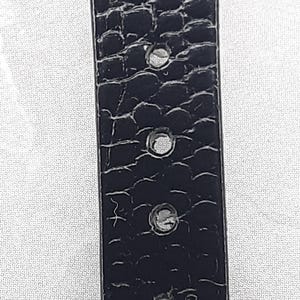 May include: Close-up of a black belt with a textured, crocodile-like pattern. The belt has several evenly spaced holes and a silver-colored buckle. The background is a white, patterned surface.