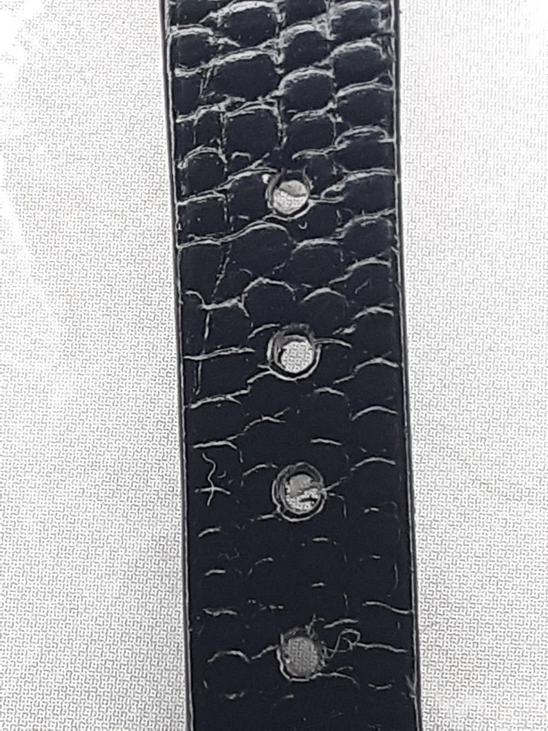 May include: Close-up of a black belt with a textured, crocodile-like pattern. The belt has several evenly spaced holes and a silver-colored buckle. The background is a white, patterned surface.
