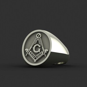 May include: A silver ring with a round, engraved design featuring a compass and the letter "G".