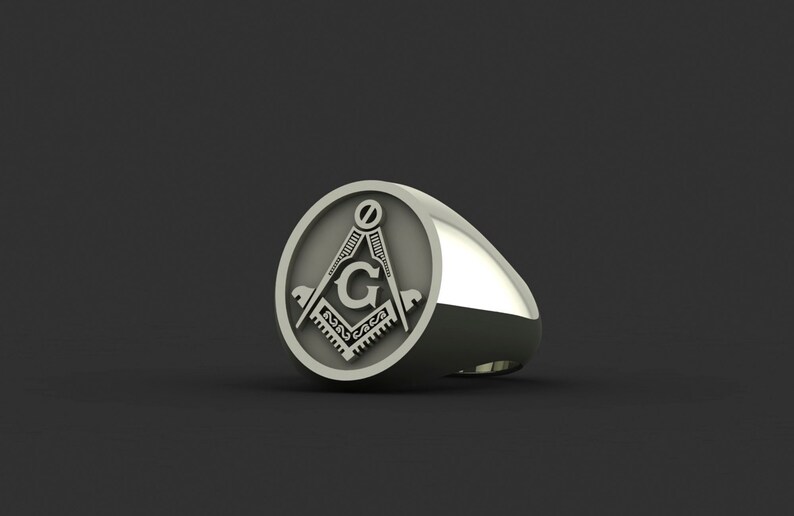 May include: A silver ring with a round, engraved design featuring a compass and the letter "G".