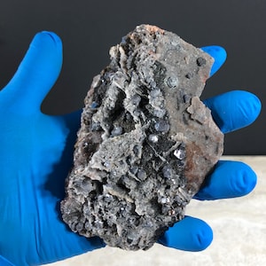 May include: A rough, irregularly shaped rock formation with a mix of gray, black, and silver metallic minerals. The rock has a textured surface with visible crystal formations and a hint of reddish-brown coloration.
