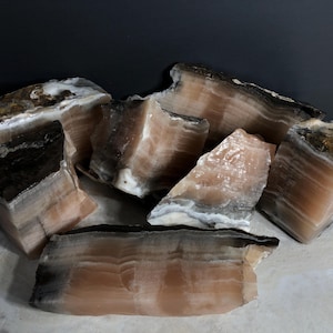May include: A collection of raw, natural calcite stone pieces. The stones display layers of tan, brown, and white hues, with a rough, textured surface. The shapes are irregular, showcasing the natural formations of the mineral.