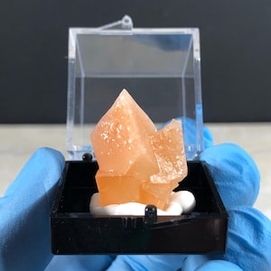 May include: A translucent, peach-colored mineral crystal cluster with sharp, angular points. The specimen is mounted on a white base inside a small, square, clear plastic display box with a black bottom, showcasing its natural geometric structure.