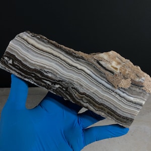 May include: A rectangular slab of banded stone featuring alternating layers of white, cream, gray, and dark brown colors. The surface displays a polished, smooth finish on the front face, while the top edge remains rough, textured, and unpolished.