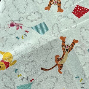 May include: A light blue fabric with a repeating pattern of Winnie the Pooh and Tigger characters. The characters are flying kites in the sky with clouds. The kites are various colors and patterns.