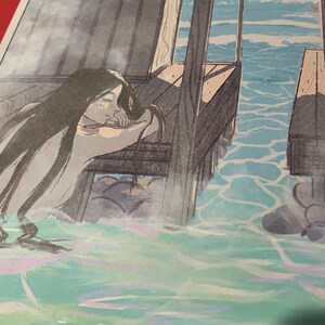 May include: A woman with long dark hair leans against a wooden railing, looking out at the ocean. The water is a light blue and green, with whitecaps. The woman's face is obscured by her hair, but she appears to be wearing a white dress.
