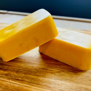 Può includere: Due saponette rettangolari gialle sono impilate su una superficie di legno. Il sapone ha un aspetto liscio, leggermente lucido e un colore giallo dorato caldo. La superficie in legno ha venature naturali.