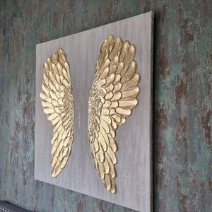 Könnte beinhalten: Ein Paar goldene Engelsflügel auf einer Leinwand mit strukturiertem Hintergrund. Die Flügel sind aus einem 3D-Material gefertigt und wirken realistisch.