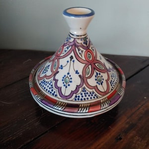 May include: A white ceramic tagine with a colorful floral design in shades of pink, blue, and orange. The tagine has a round base and a conical lid.