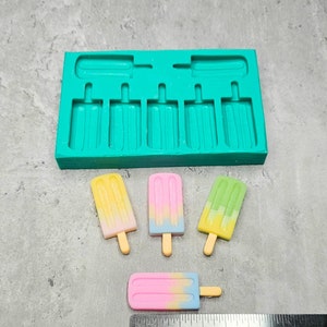Może przedstawiać: Forma silikonowa z sześcioma kształtami lodów na patyku i czterema kolorowymi cukierkami w kształcie lodów na patyku. Cukierki są żółte, różowe i zielone. Dołączono linijkę do odniesienia rozmiaru.