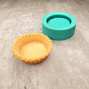 May include: A close-up of a small, round, yellow pastry tart shell. The tart shell is sitting next to a green silicone mold with the words "Cotton Ar" visible on the side.