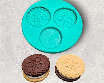 Puede incluir: Un molde de silicona turquesa con dos impresiones redondas de un diseño de galleta y una impresión redonda más pequeña en el centro. Dos galletas, una con la parte superior marrón y otra con la parte superior beige, se muestran junto al molde.