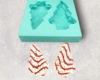 May include: A teal silicone mould with two Christmas tree shapes. Two decorated Christmas tree cookies are shown below the mould. The cookies are decorated with white icing, red stripes, and green sprinkles.