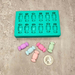 Op de afbeelding: Een groene siliconen mal met 16 holtes in de vorm van snoepjes. Vijf kleine snoepjes in de vorm van snoepjes worden getoond in verschillende pastelkleuren: lichtblauw, roze, lichtgroen en paars.