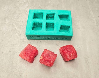 May include: A teal blue silicone mold with six square cavities. The mold is labeled "Vanilla Cotton Aromas". Three red cubes of watermelon are in front of the mold.