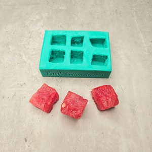 Puede incluir: Un molde de silicona azul turquesa con seis cavidades cuadradas. El molde está etiquetado como "Vanilla Cotton Aromas". Tres cubos rojos de sandía están delante del molde.