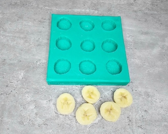 May include: A turquoise silicone mold with 12 round cavities, perfect for making small treats or desserts. Five slices of banana are arranged below the mold.