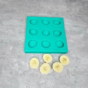 以下が含まれることがあります： 小さな御馳走やデザートを作るのに最適な、12個の丸い空洞を持つターコイズブルーのシリコンモールド。モールドの下にバナナのスライスが5枚並んでいます。