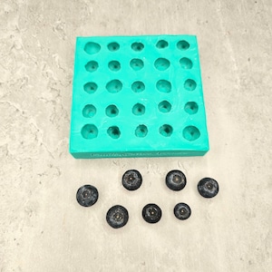 Puede incluir: Un molde de silicona turquesa con 36 cavidades redondas, diseñado para hacer objetos pequeños y redondos. El molde está sobre una superficie gris. Hay siete objetos pequeños de color azul oscuro dispersos alrededor del molde.